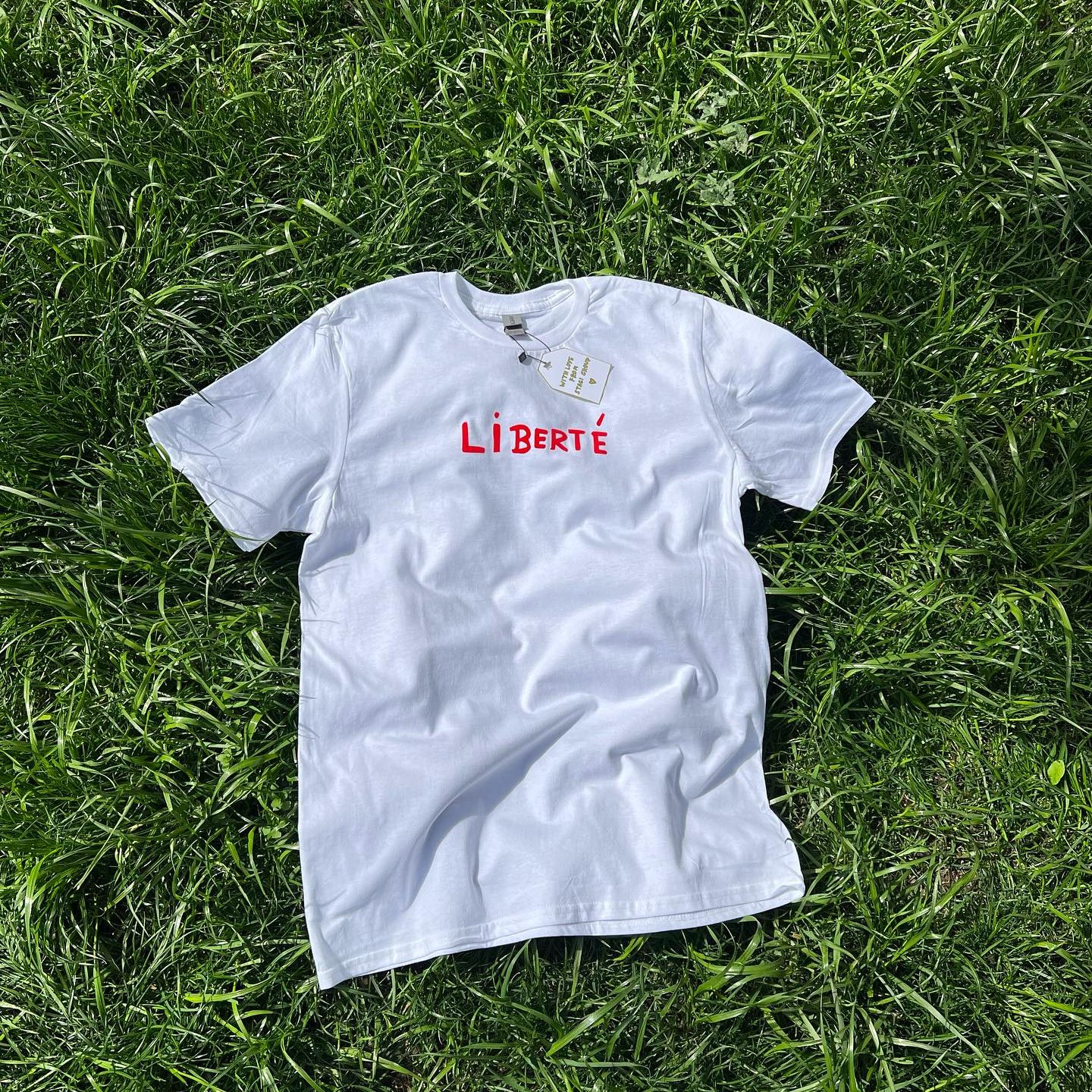 Stagi Group, სტაგი ჯგუფის თეთრი მაისური, წითელი წარწერა, LIBERTE, ხავერდის დიზაინი, ბალახის ფონზე