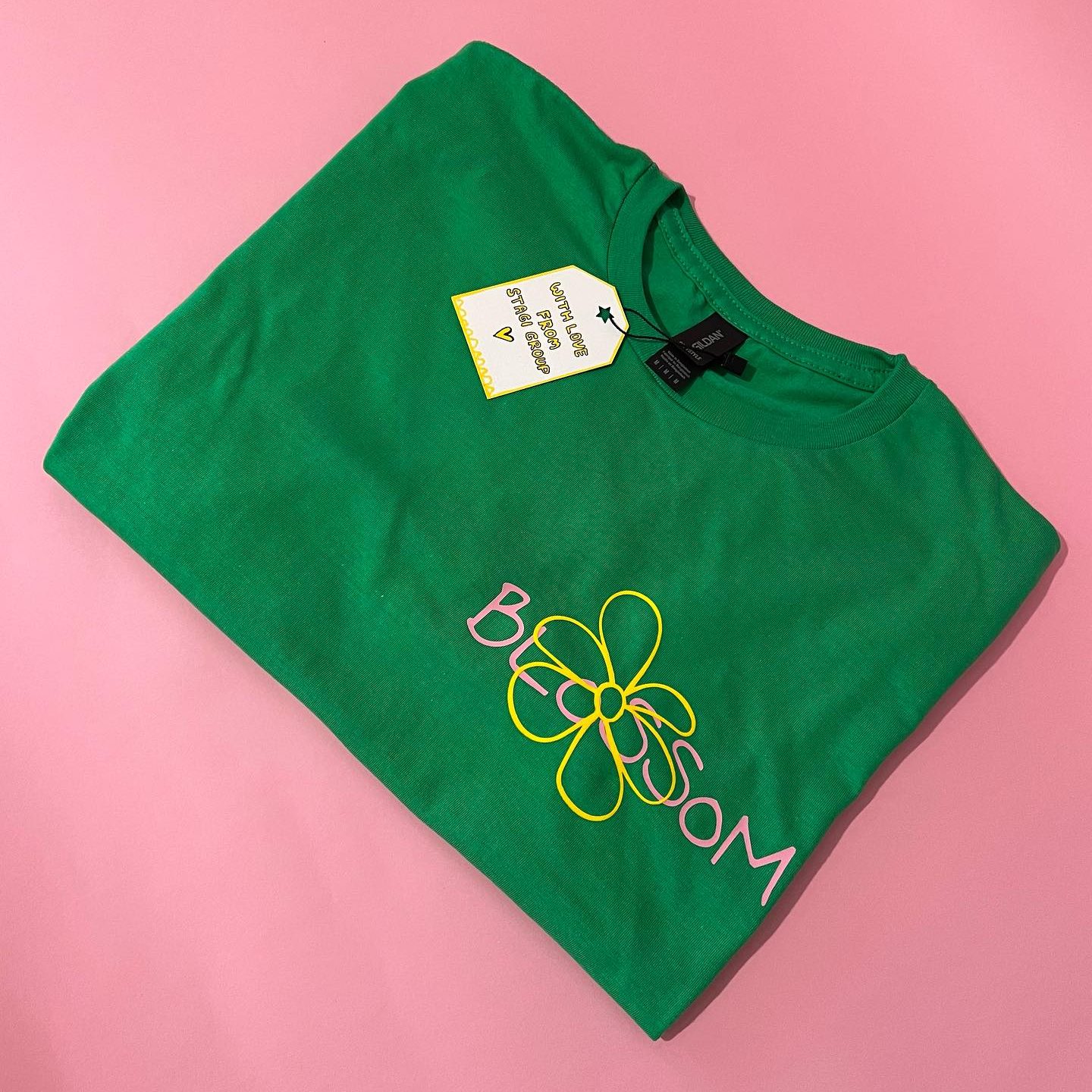 Stagi Group, სტაგი ჯგუფის მწვანე მაისური, ვარდისფერი წარწერა, BLOSSOM, ყვავილის ყვითელი დიზაინით, ვარდისფერ ფონზე