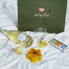 პრემიუმ სასაჩუქრე ნაკრები მწვანე და კრემისფერი ხავერდით, შამპანიურის ბოთლით, ორი ჭიქით და შოკოლადის ნაკრებებით, A green velvet gift box closed and displayed on white fabric, with its contents, including a Prosecco bottle, flower-shaped container, heart-shaped chocolates, and decorative glasses, neatly arranged around it.